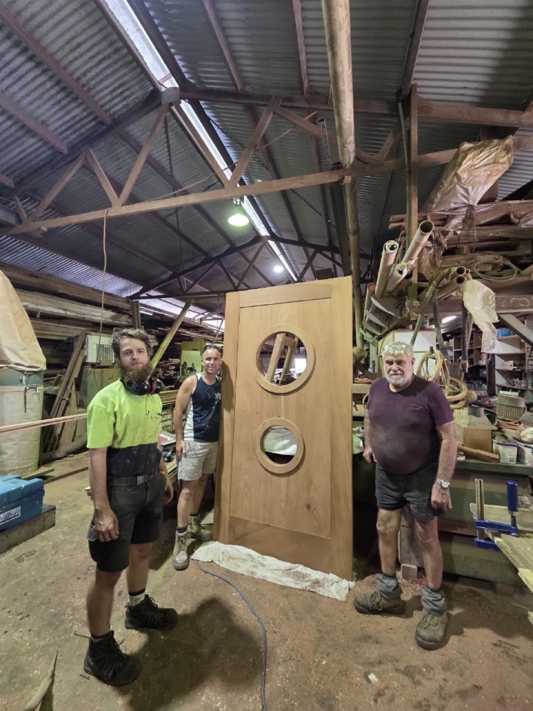 Three cabinet makers standing beside a tall timber door with two round glazed portholes in the workshop