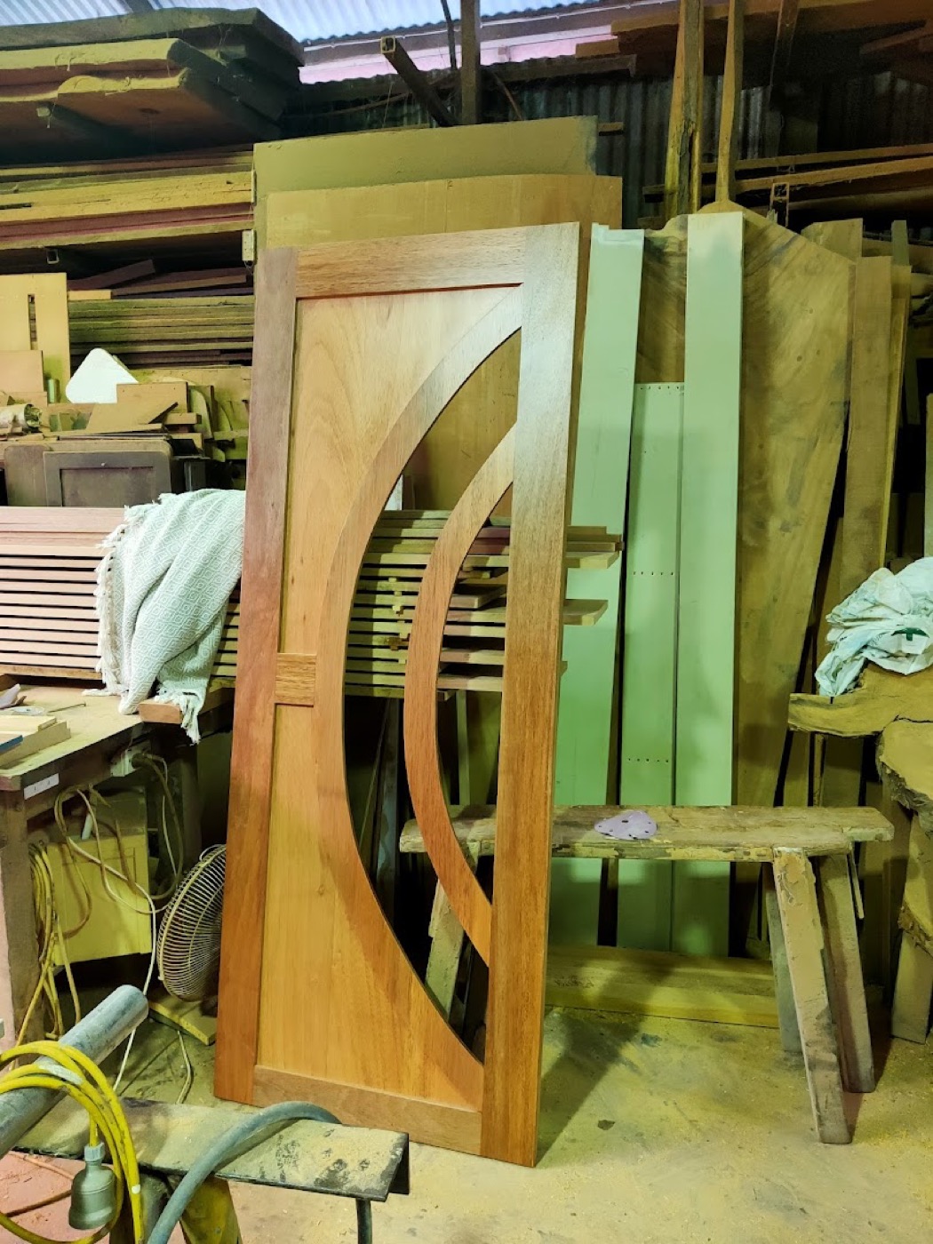 A curved timber component clamped upright beside stacked boards in the workshop