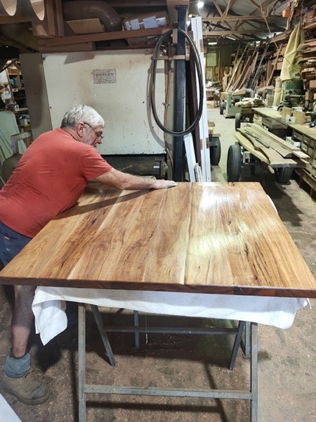 A cabinet maker leaning over a large solid timber table top, hand-sanding the surface