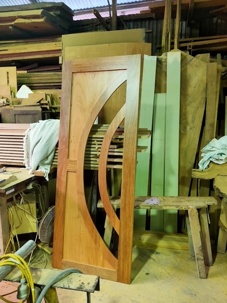 A curved timber component standing upright beside stacked boards in the workshop